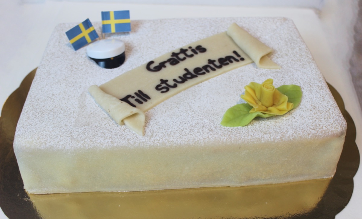 Klassisk studenttårta som en vit studentmössa med blågult band, gratulations text och vackra blommor